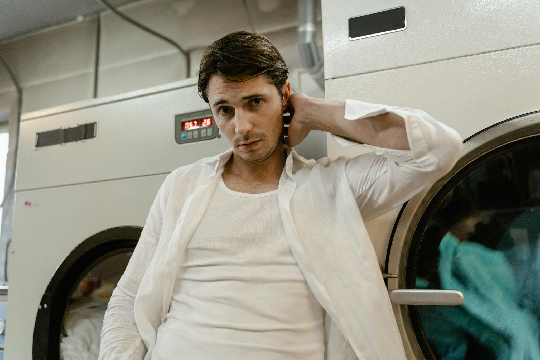 Young man in a white shirt leaning on a washing machine in a laundry facility.